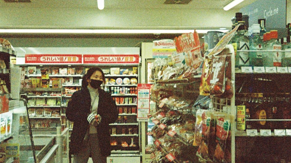 戴口罩的顧客站在店內走道，一旁掛架擺滿各式零食袋，後方貨架掛著日文紅色價格標牌